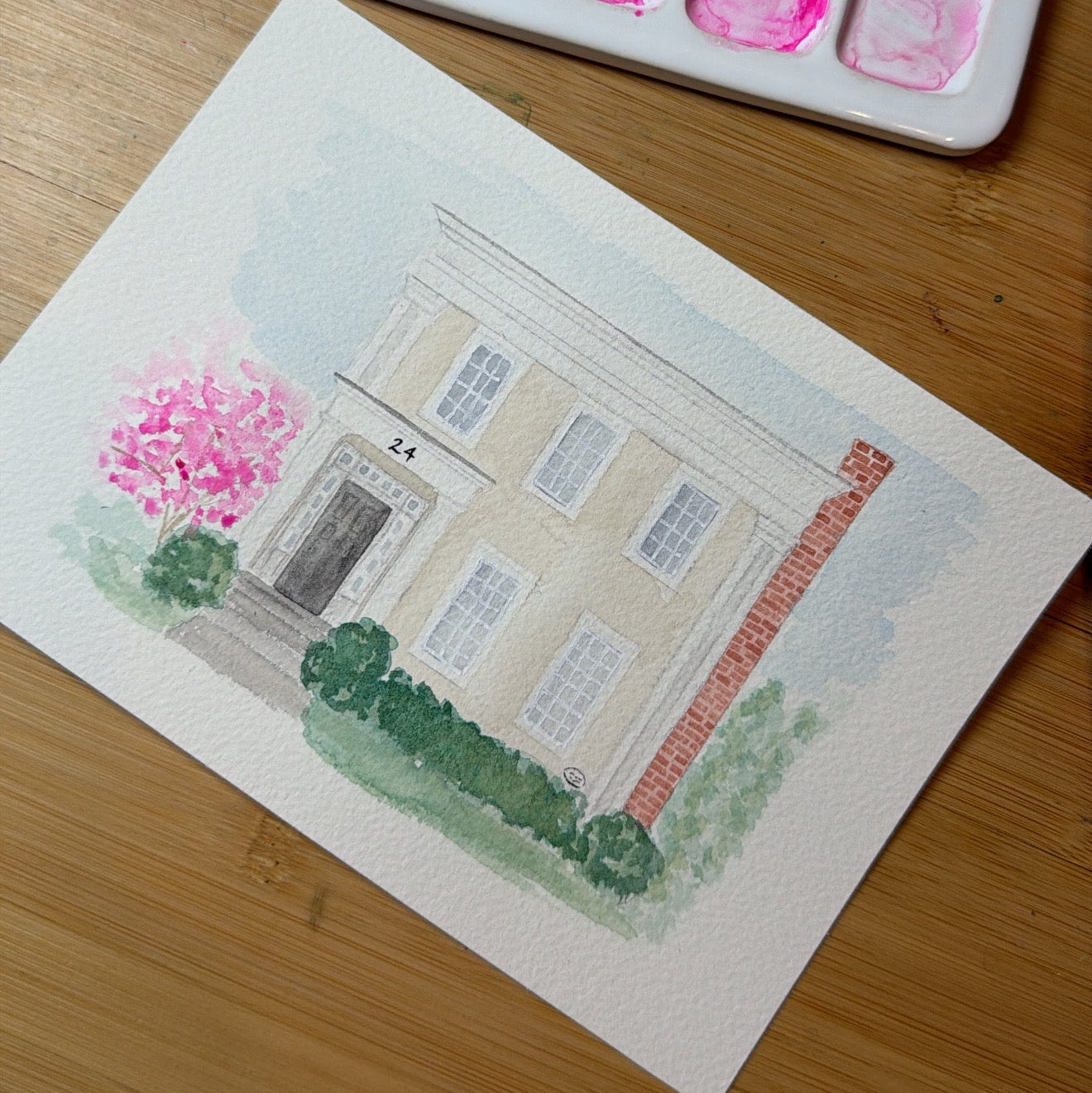 Watercolor painting of a house on paper with a watercolor palette in the background.