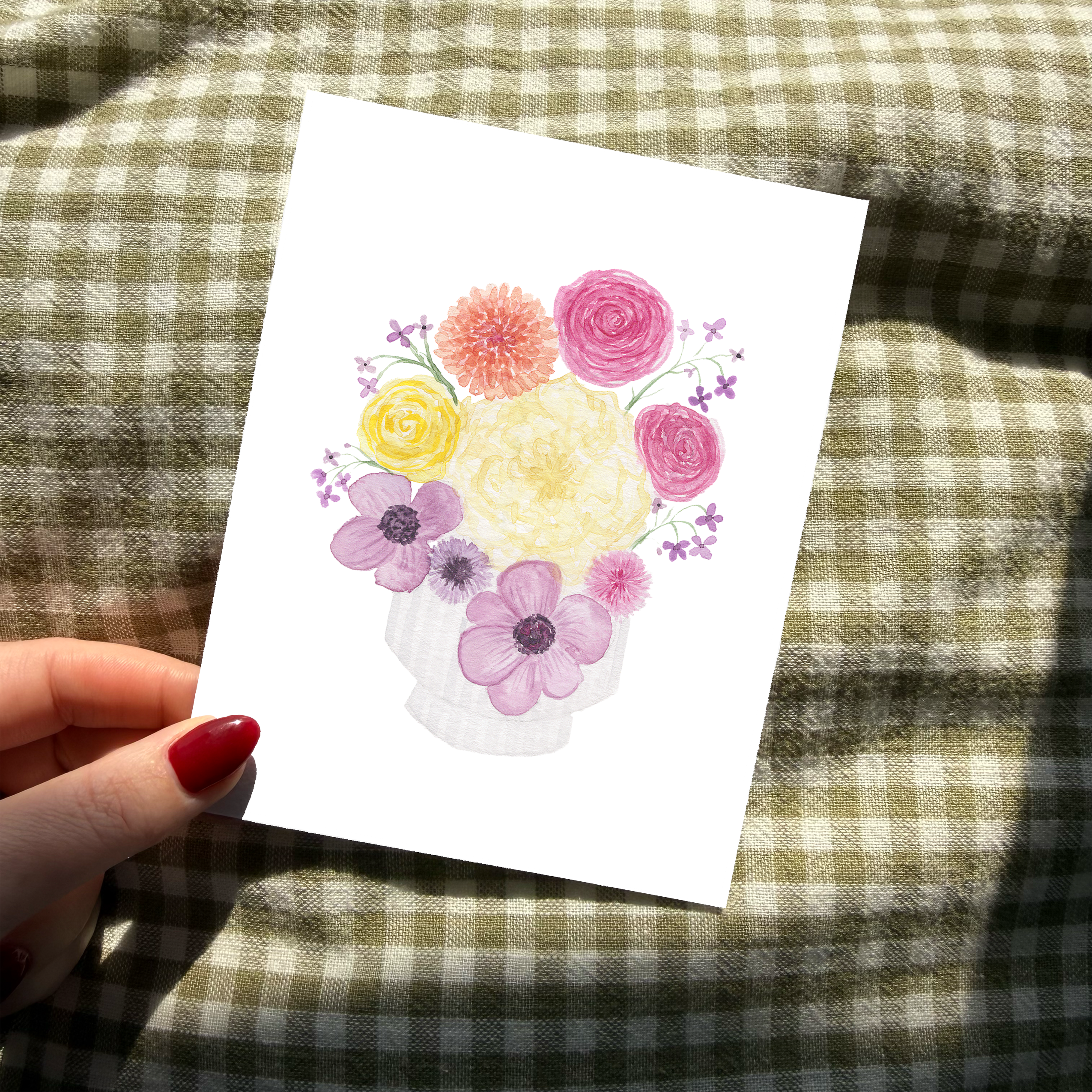 A hand with a red nail is holding a card with a flower bouquet in a jar painted on it with a green  and white checkered background.