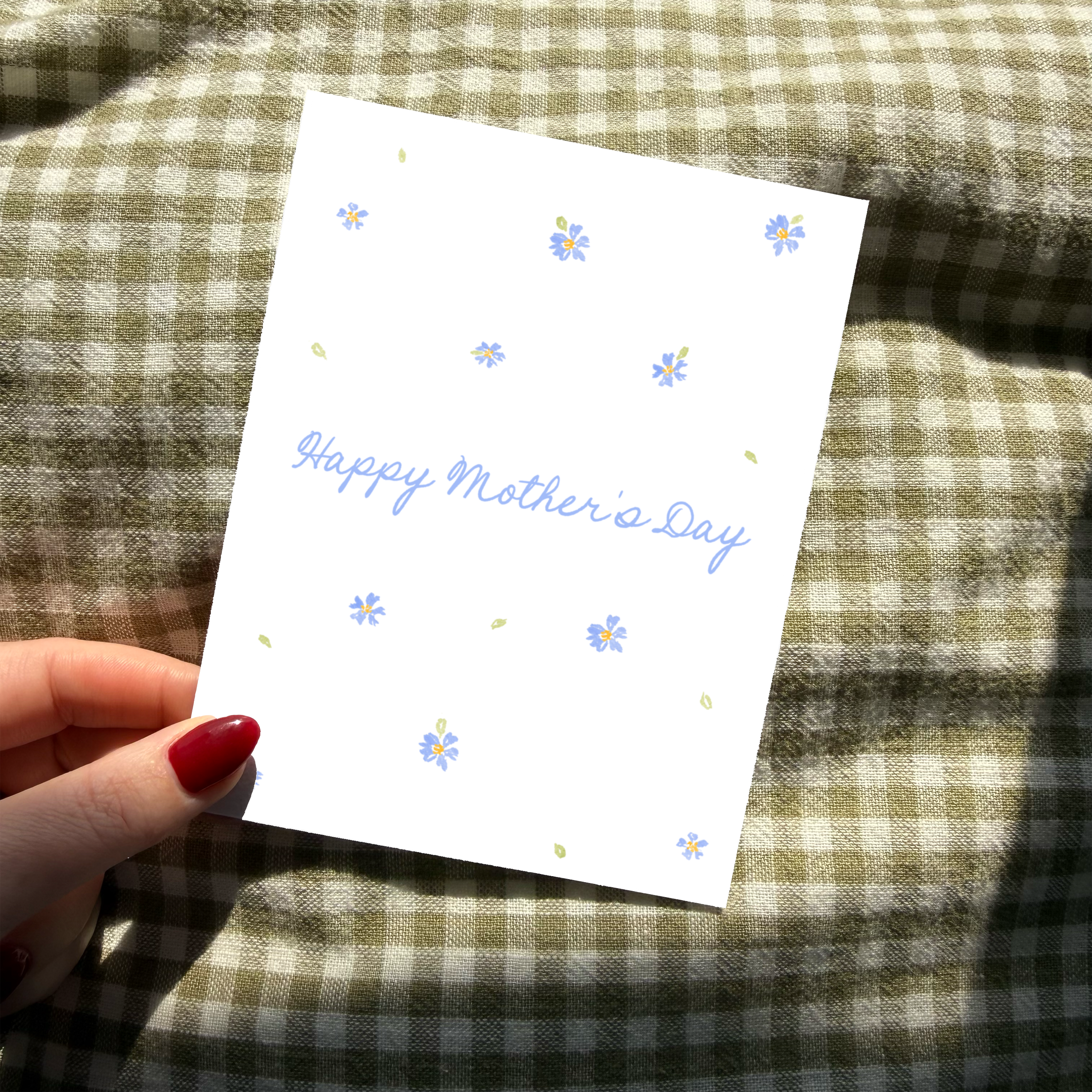 Hand holding a 'Happy Mother's Day' card with floral designs on a checkered fabric background