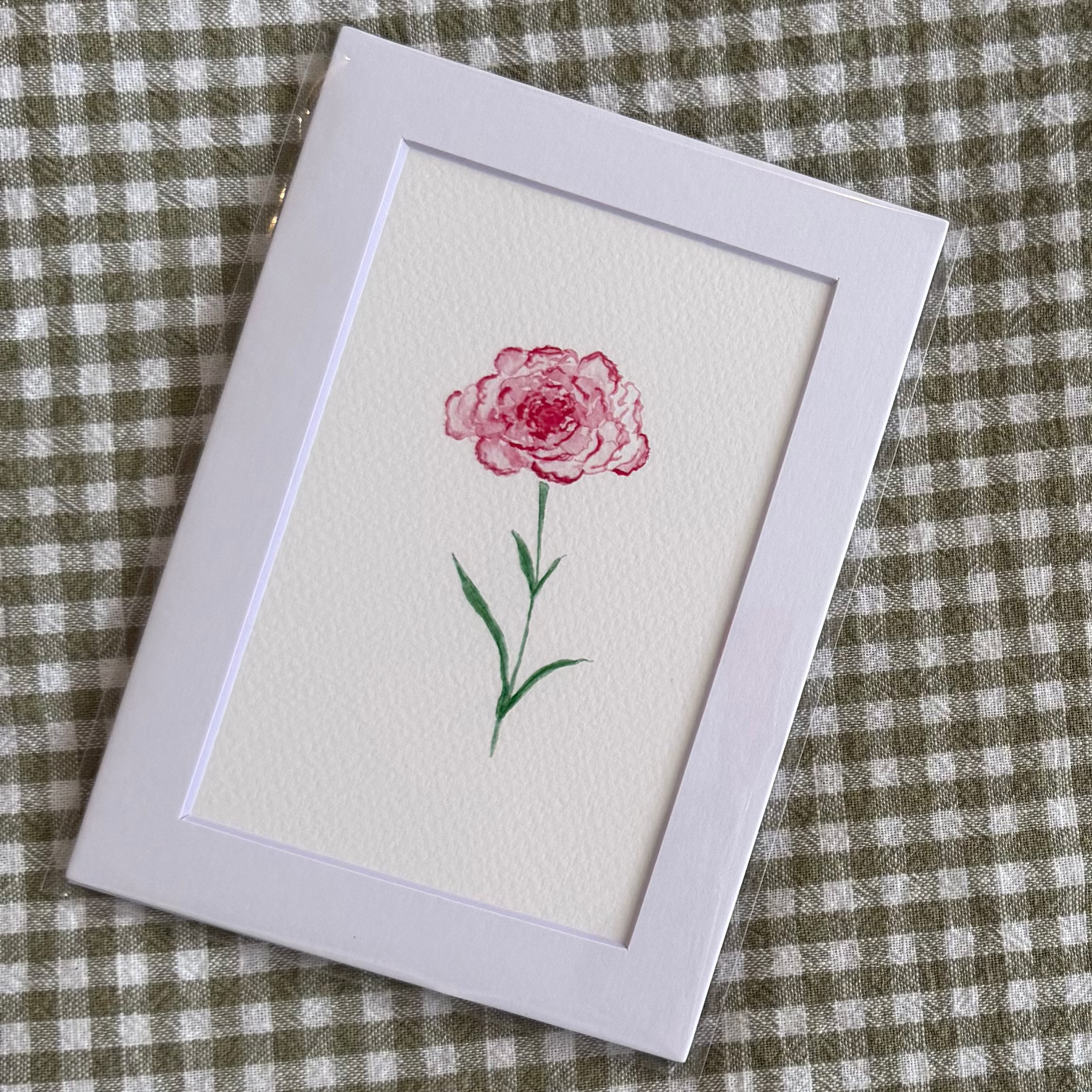 Small framed artwork of a pink carnation flower on a green and white checkered fabric background