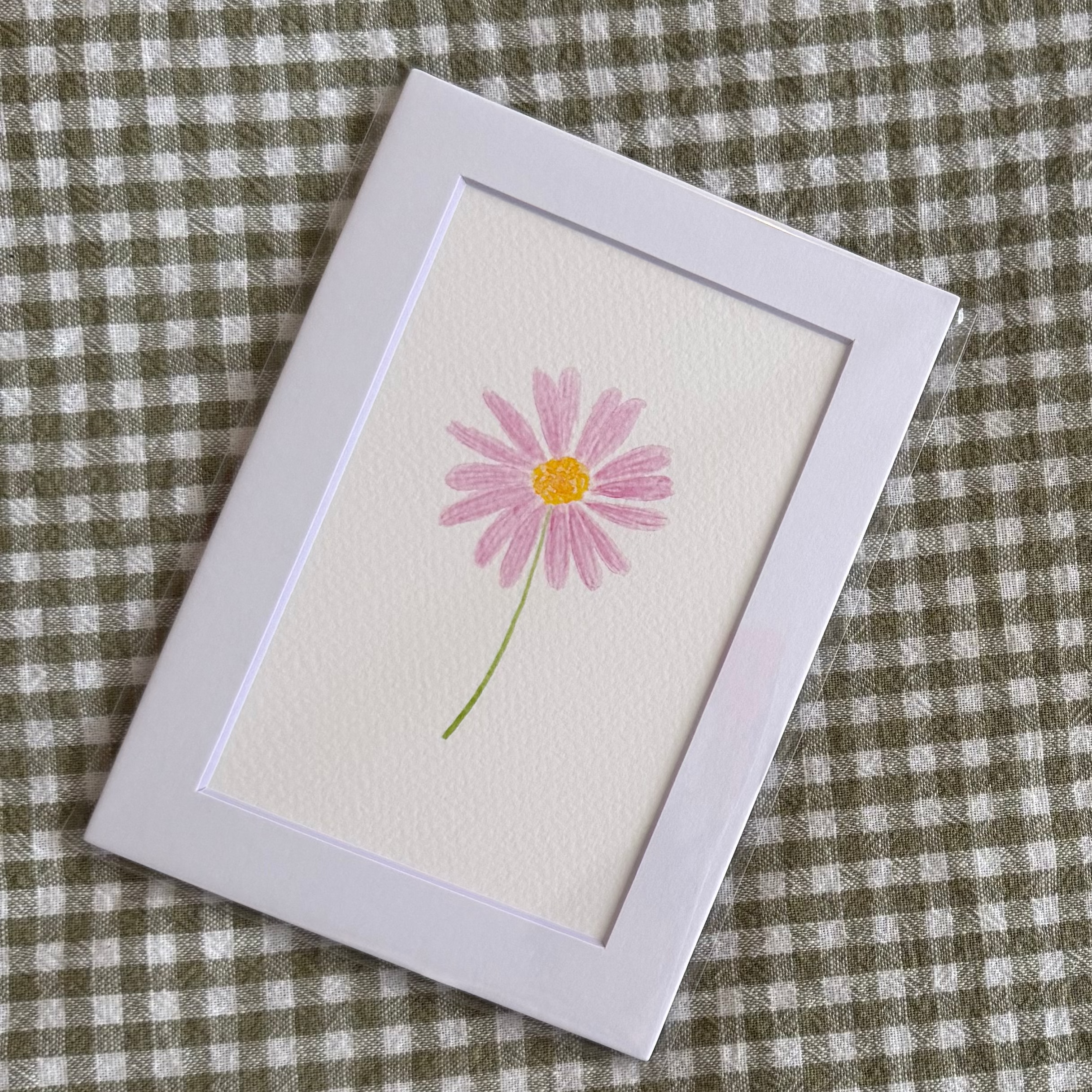 Small framed picture of a pink daisy flower on a green and white checkered fabric background