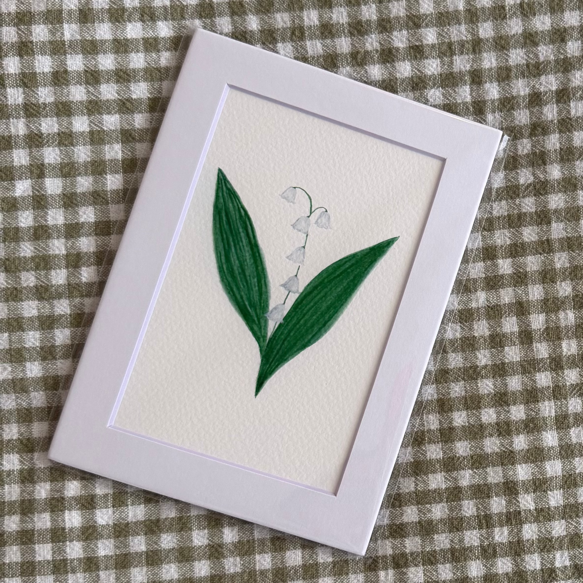 Small framed artwork with a lily of the valley painting on a green and white checkered fabric background