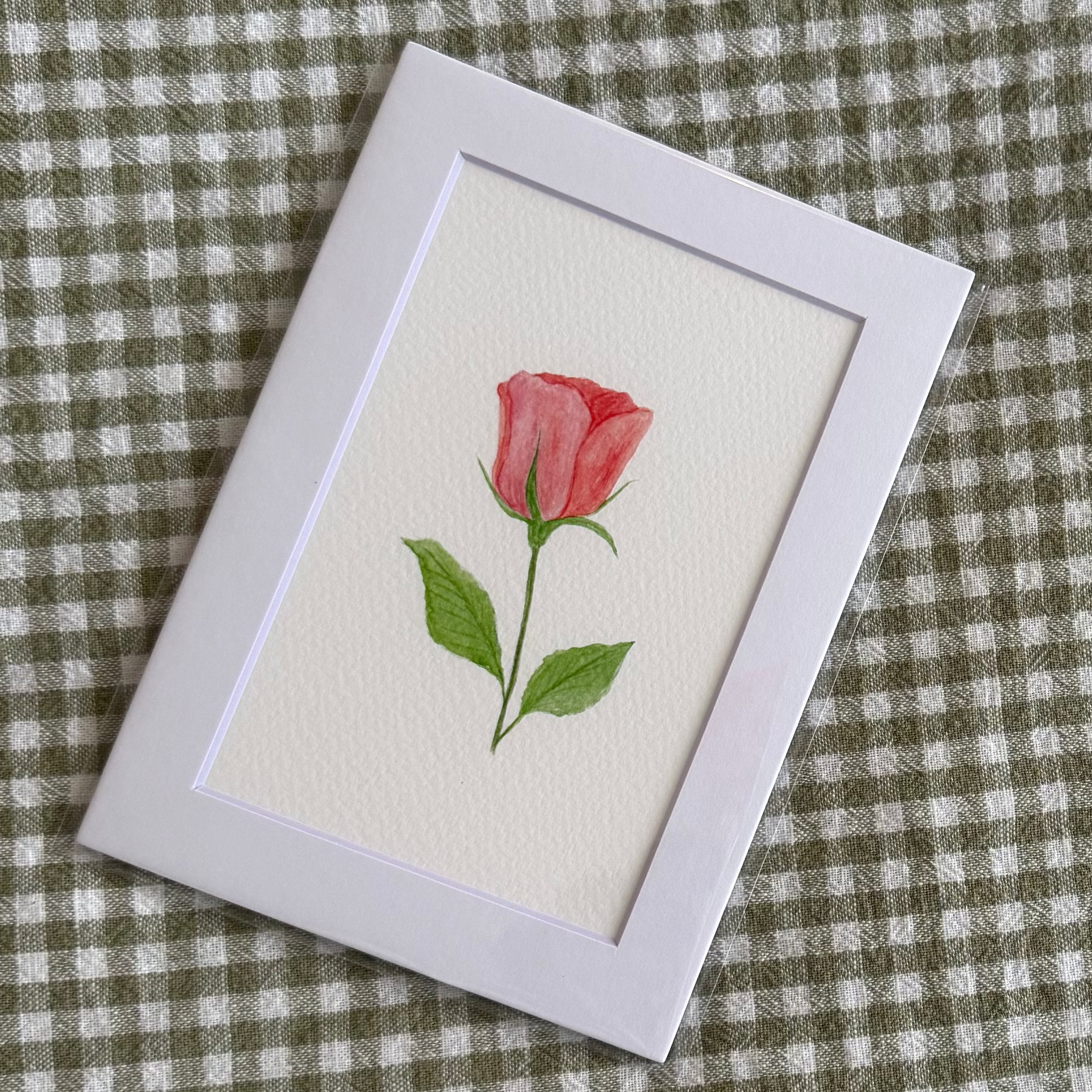 Small framed artwork of a pink rose on a green and white checkered fabric background