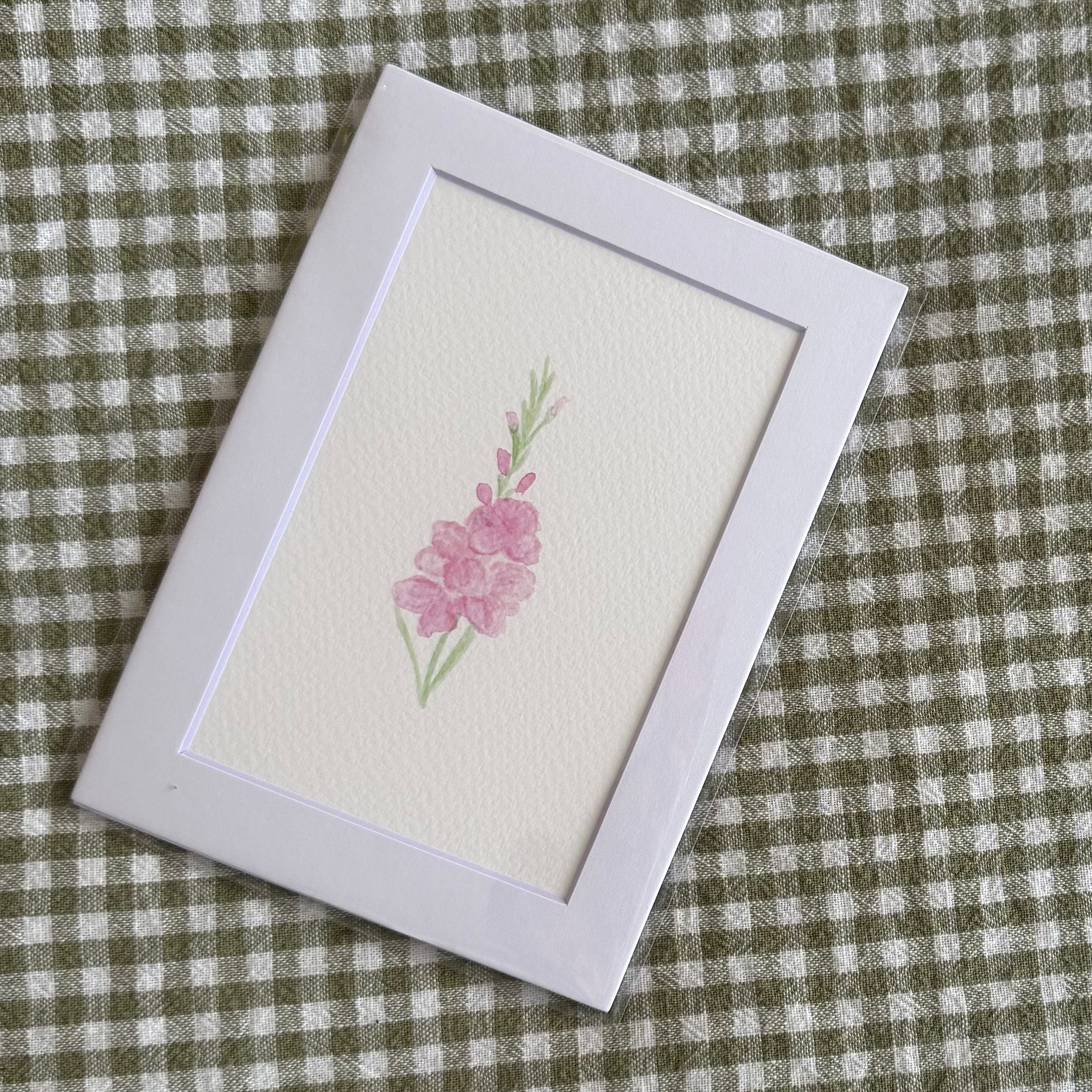Small framed artwork with a pink flower on a green and white checkered fabric background
