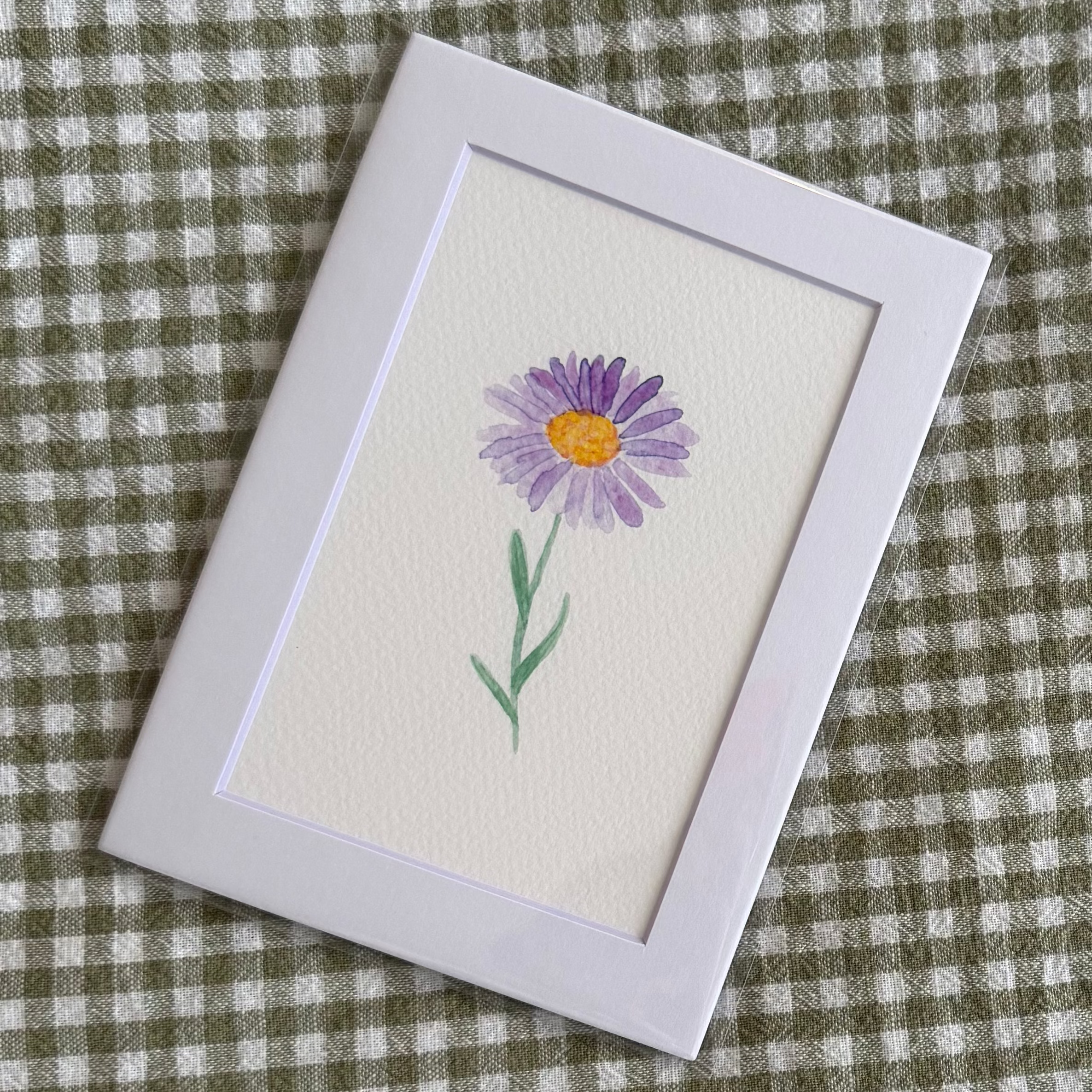 Small framed artwork of a purple aster flower on a green and white checkered fabric background