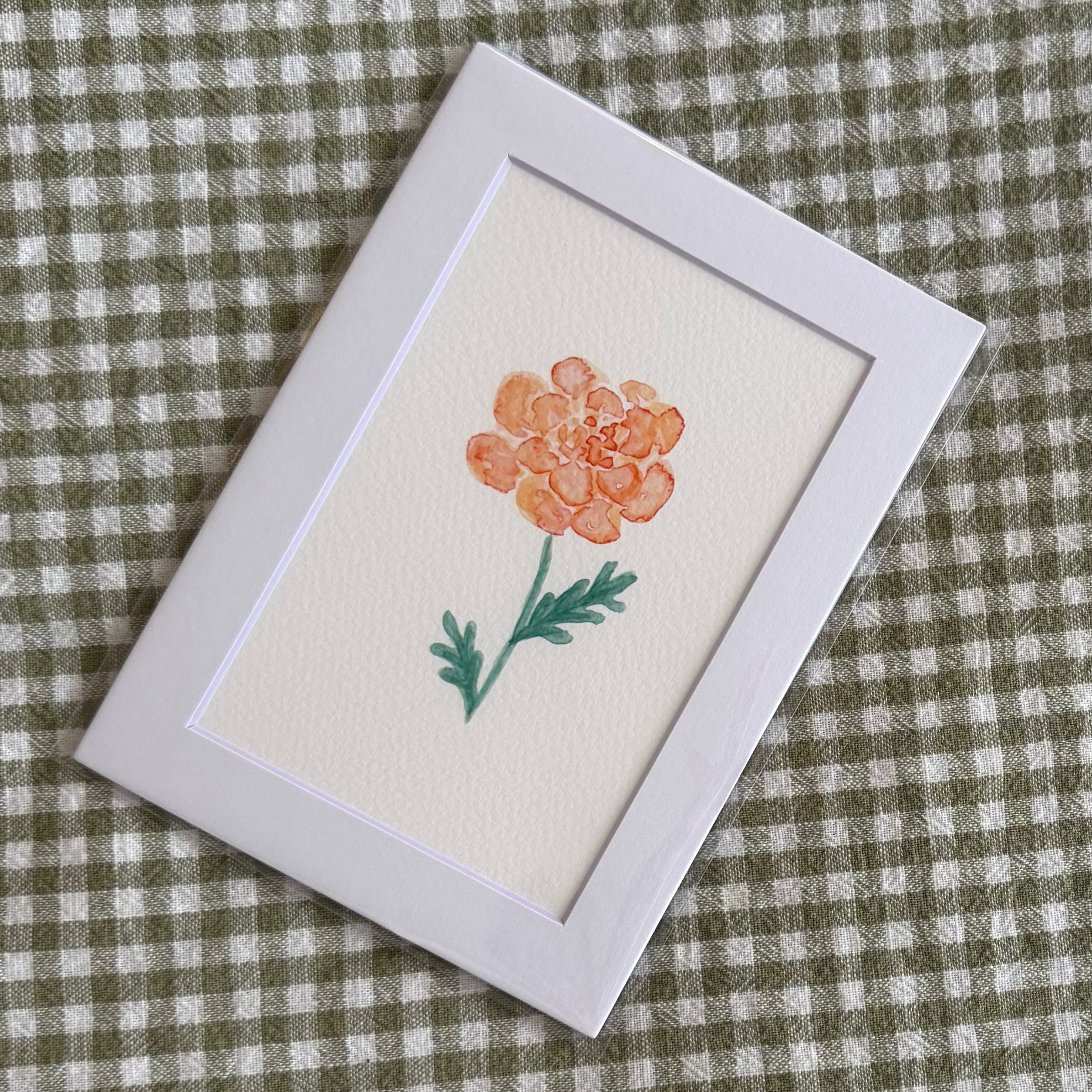 Small framed artwork of a marigold flower on a green and white checkered fabric background