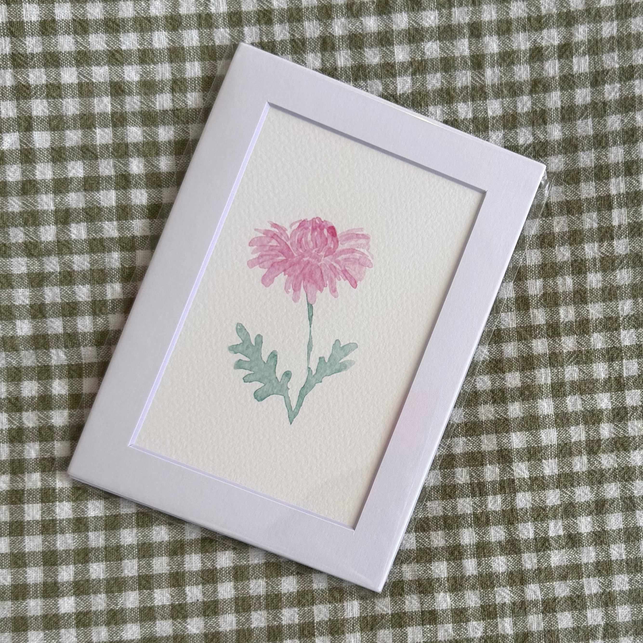 Small framed artwork of a pink chrysanthemum flower on a green and white checkered fabric background
