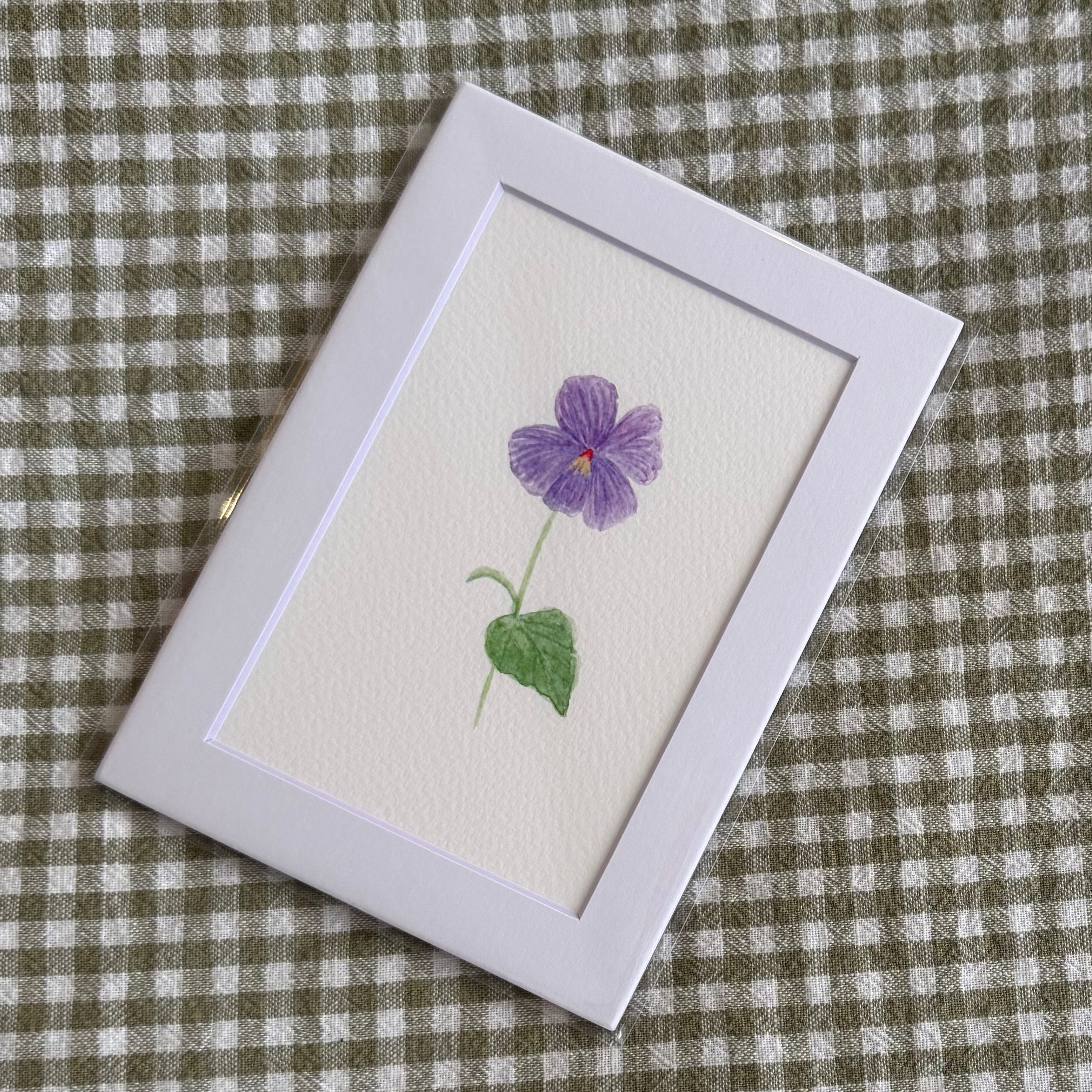 Small white framed art print with a purple violet flower on a green and white checkered fabric background