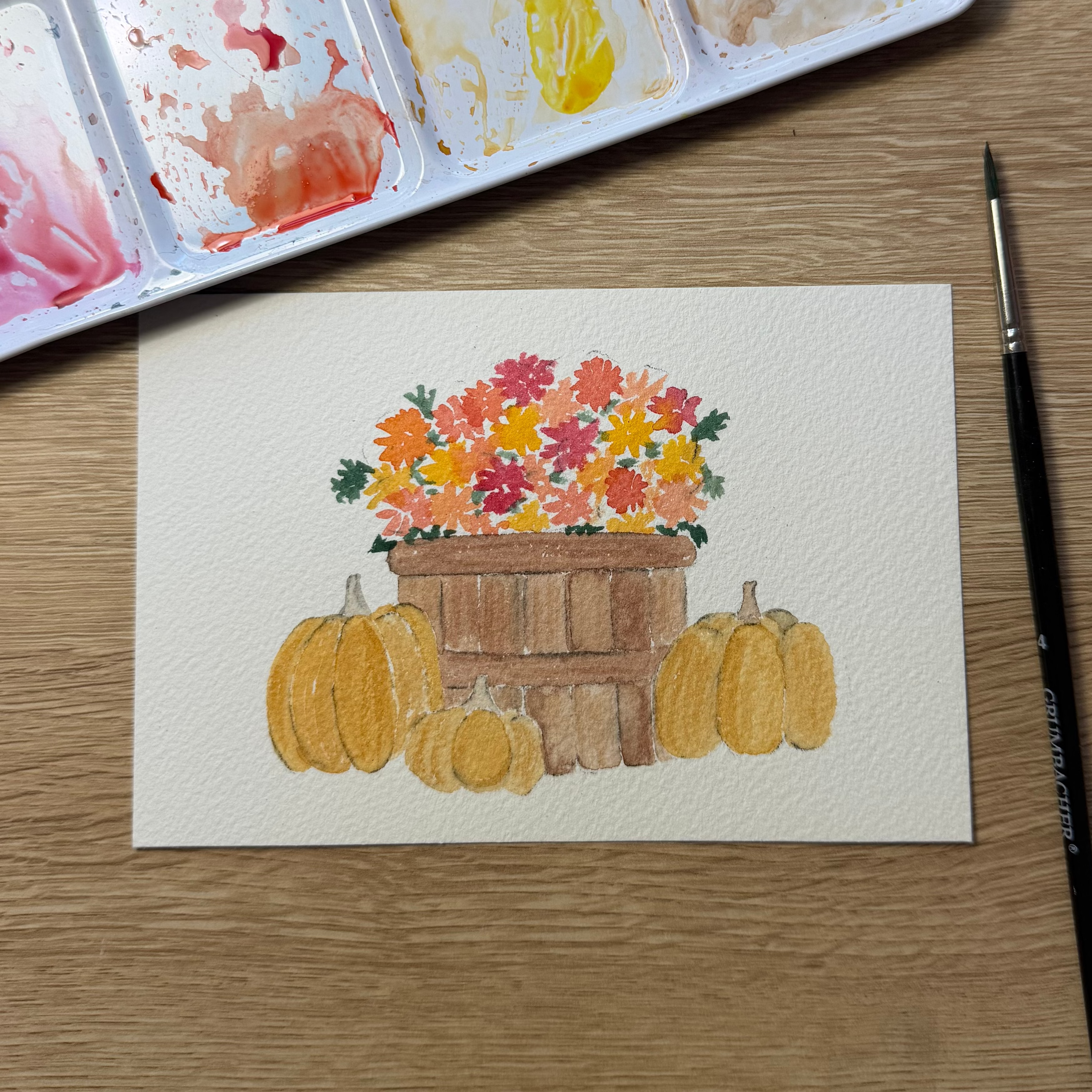 A watercolor painting of pumpkins and flowers in a basket, displayed on a wooden table next to a watercolor palette and paintbrush.