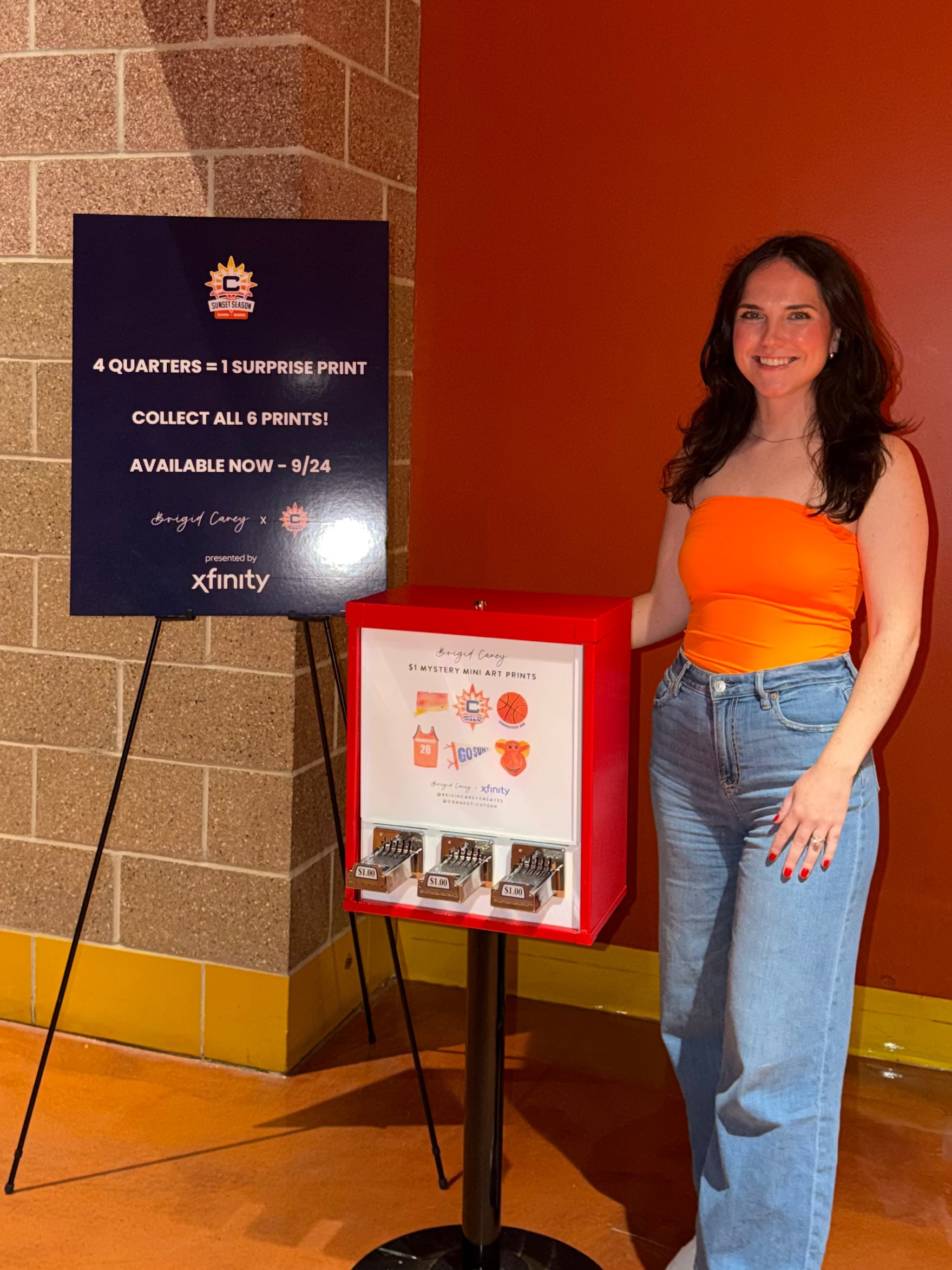 A woman stands next to a red and white display stand featuring a custom Connecticut Sun mini watercolor art prints, sponsored by Xfinity, set in an indoor venue with orange walls and a brick column.