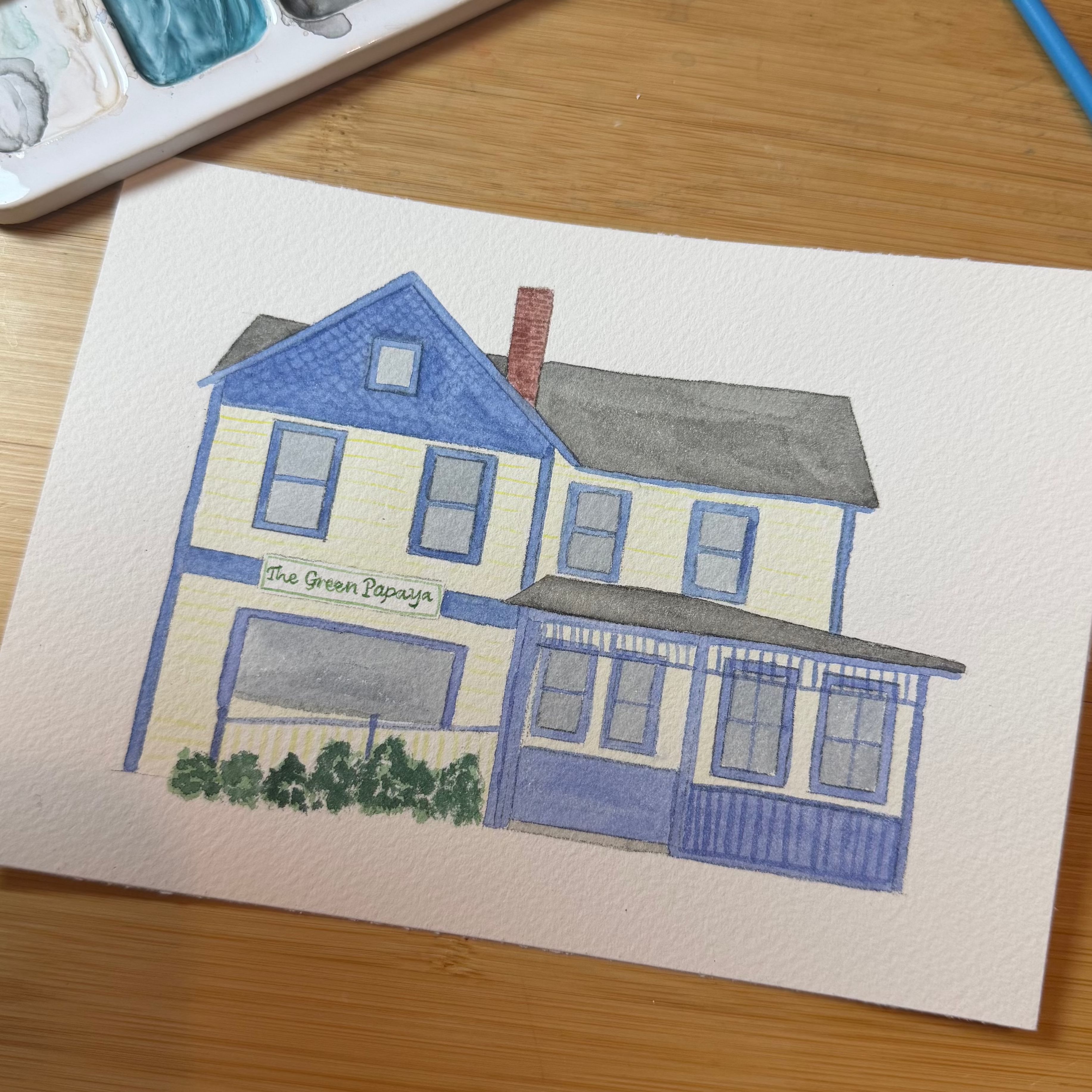 Painting of a house on paper with watercolor palette and pencils on a wooden surface