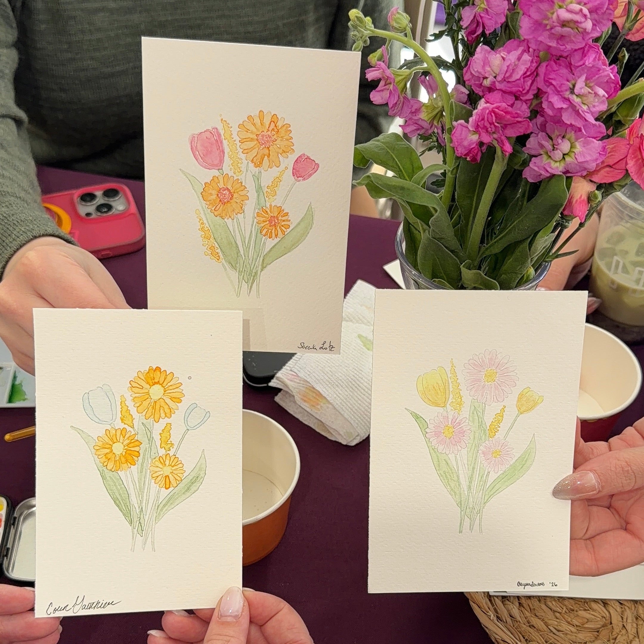Three watercolor paintings of flowers held by hands on a table with a floral arrangement.