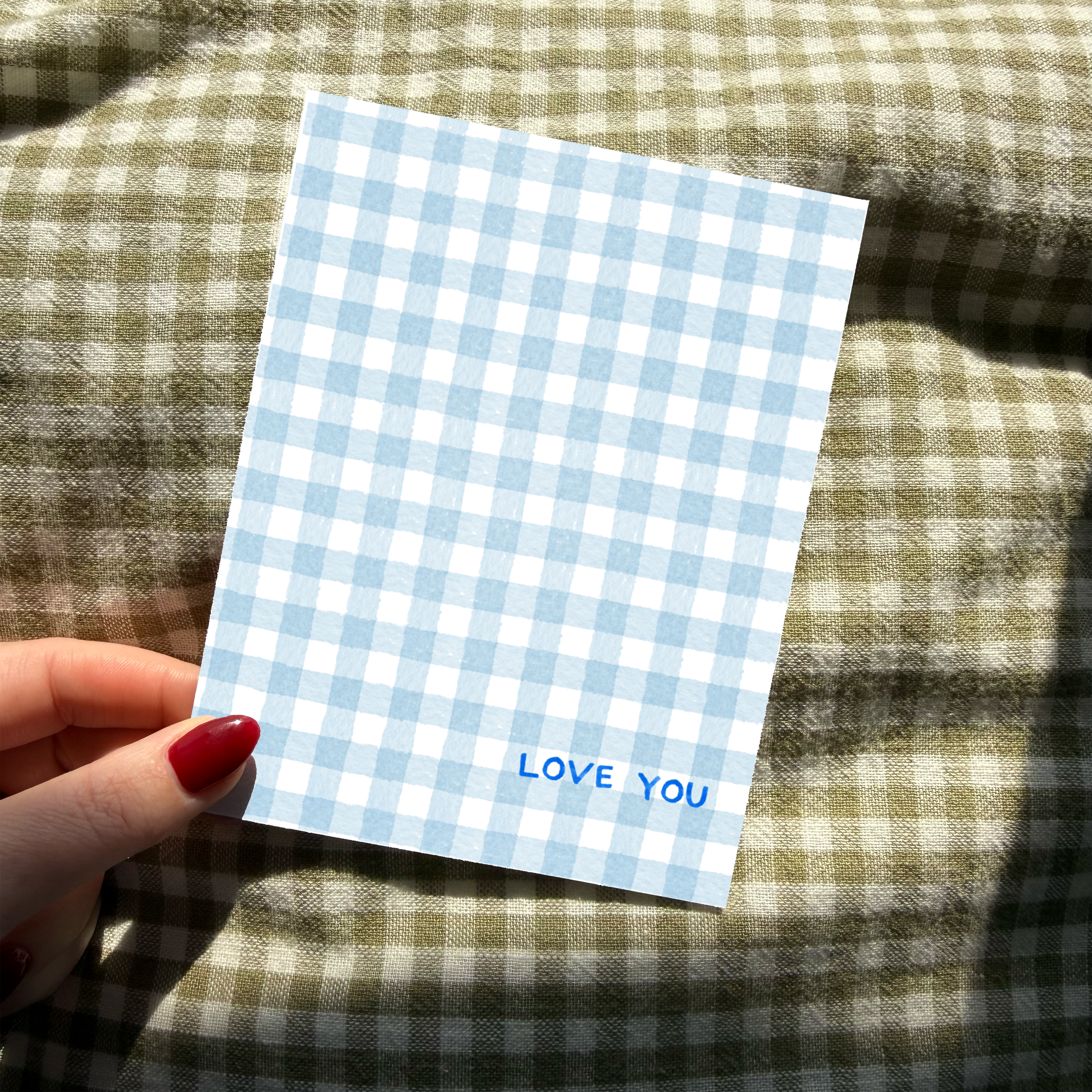 Hand holding a blue and white checkered card with 'LOVE YOU' text on a plaid fabric background